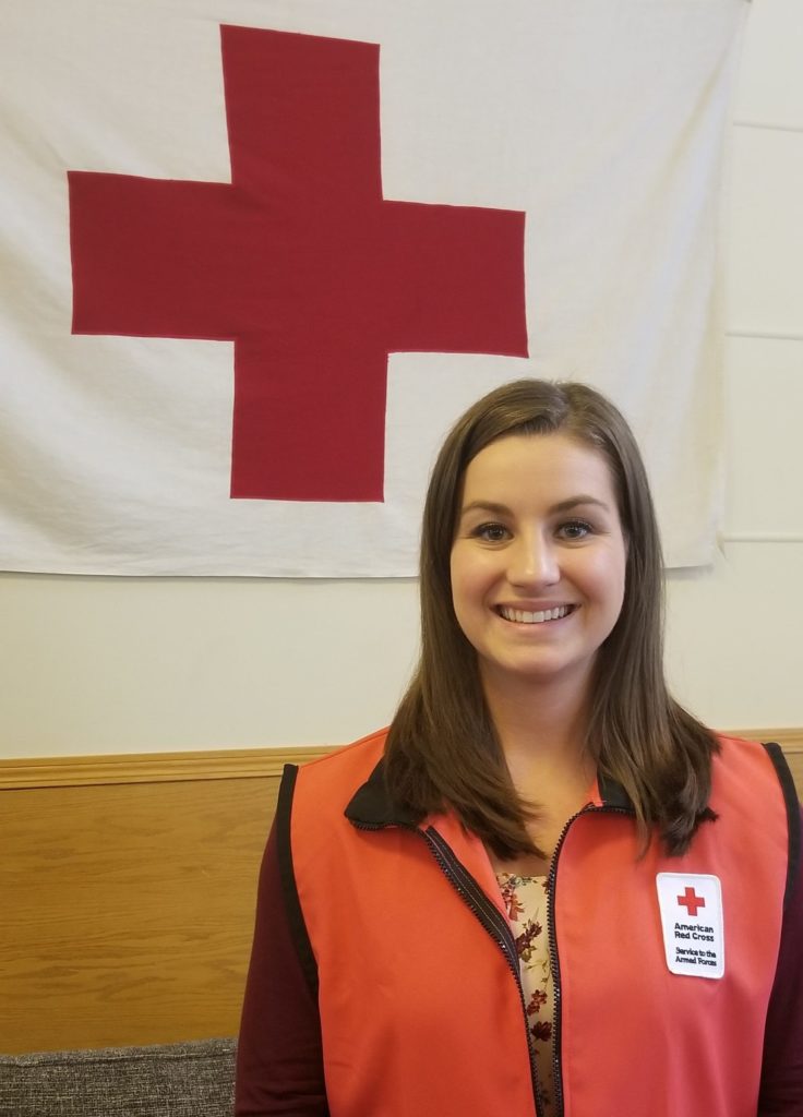 Lauren in her Red Cross vest standing in front of a Red Cross banner.