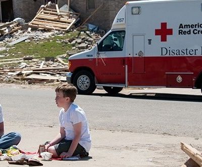Two victims of a disaster sitting on the ground near a Red Cross emergency response vehicle.