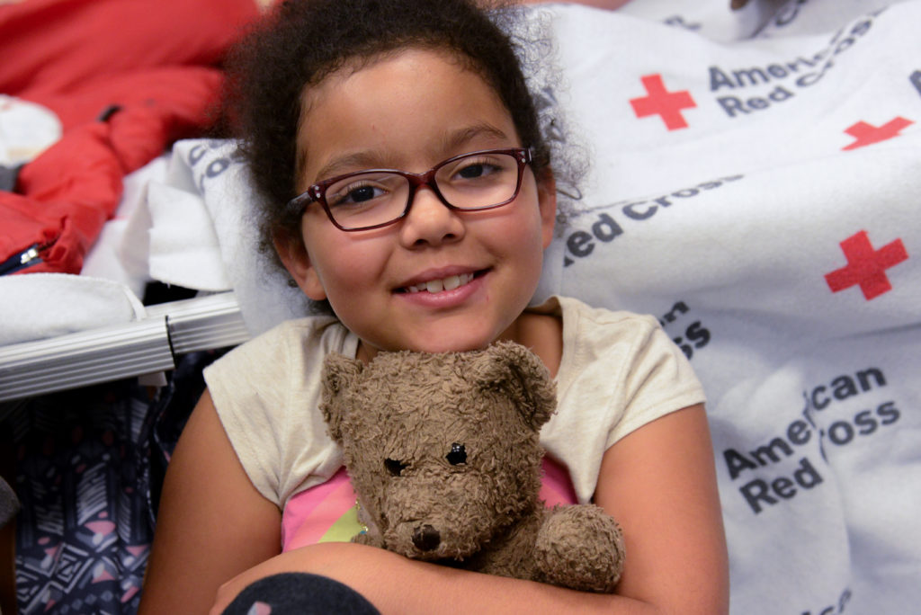 A little girl in a Red Cross shelter hugging her teddy bear. 