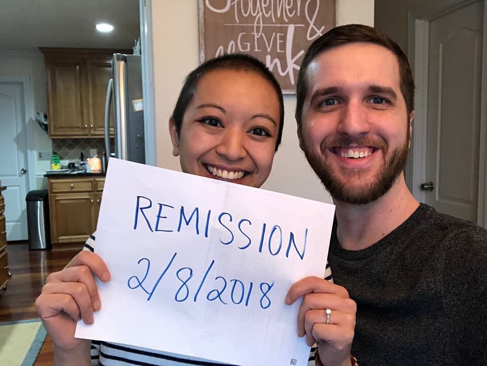 Stephanie and Justin with engagement ring and a sign about her remission.