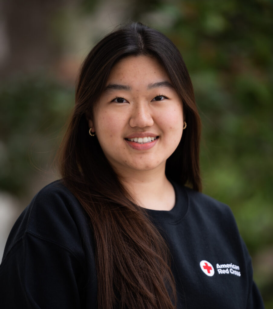 A headshot of a young woman with long dark brown hair and a long sleeve black shirt with an American Red Cross logo. 